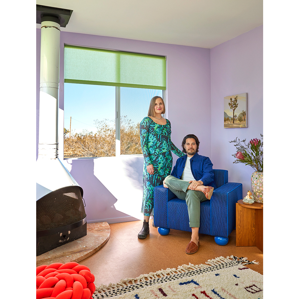 Couple poses in a modern living room with lavender walls, featuring a blue chair, fireplace, rug, and a view through the window.