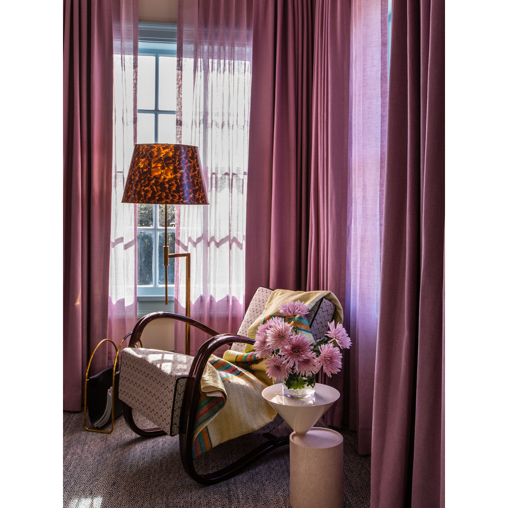 Cozy interior featuring a patterned armchair, floor lamp, side table with flowers, and mauve curtains in front of a window.