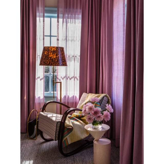 Cozy interior featuring a patterned armchair, floor lamp, side table with flowers, and mauve curtains in front of a window.