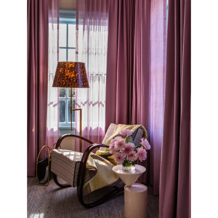 Cozy interior featuring a patterned armchair, floor lamp, side table with flowers, and mauve curtains in front of a window.