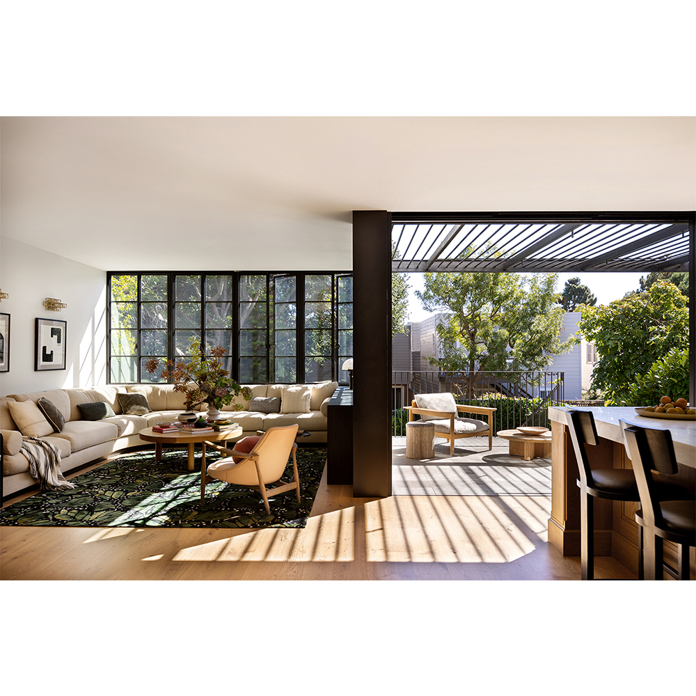 Bright modern home interior featuring a large beige sectional sofa, round wood coffee table, and floor-to-ceiling windows overlooking trees.