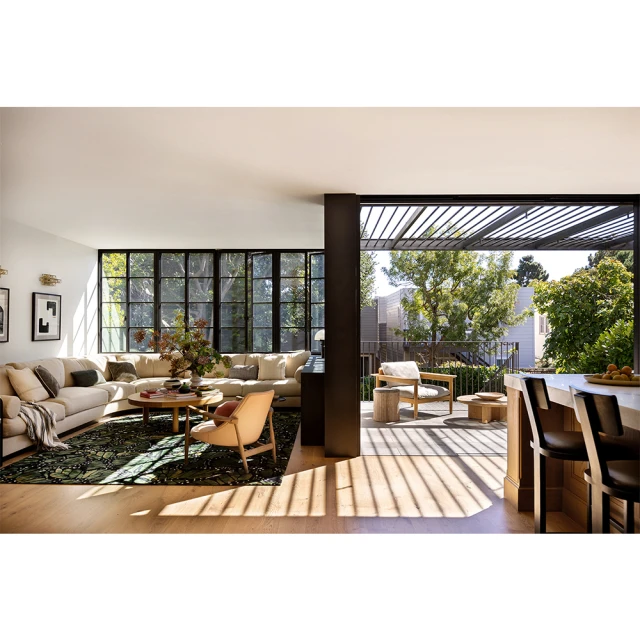 Bright modern home interior featuring a large beige sectional sofa, round wood coffee table, and floor-to-ceiling windows overlooking trees.