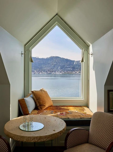 Window seat with tan cushions, pillows, blanket, and a round side table overlooking a town nestled in the hills by a body of water.