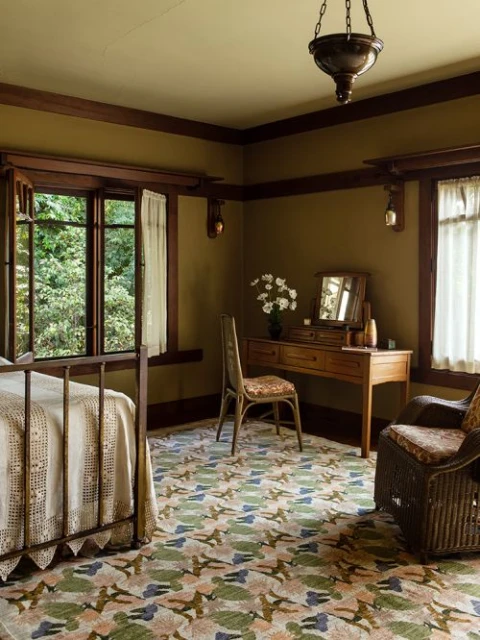 Bedroom interior featuring a bed, dressing table with a mirror, floral patterned rug, wicker chair, and natural light streaming through windows.