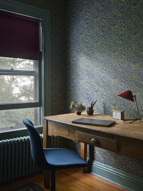 Interior scene with a blue chair in front of a wooden desk, a window with dark-red shade, and a wallpapered wall in blue and light-green.