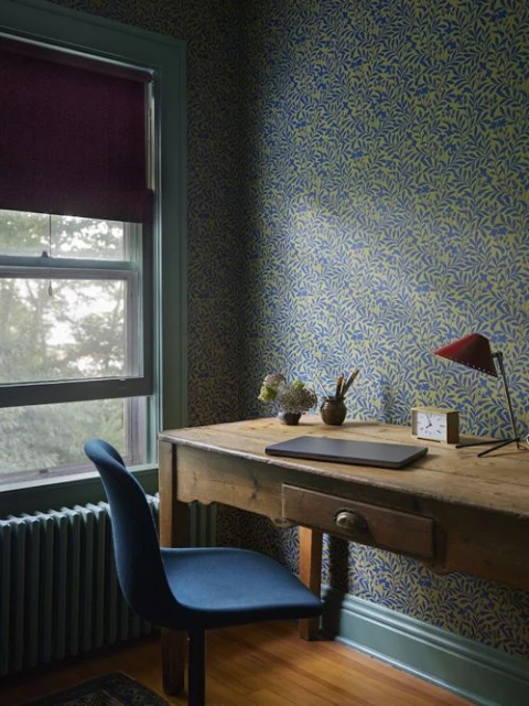 Interior scene with a blue chair in front of a wooden desk, a window with dark-red shade, and a wallpapered wall in blue and light-green.