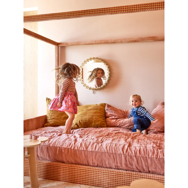 Two young children, a girl jumping on a bed with a pink bedspread and pillows, inside a bedroom with a round mirror on the wall.