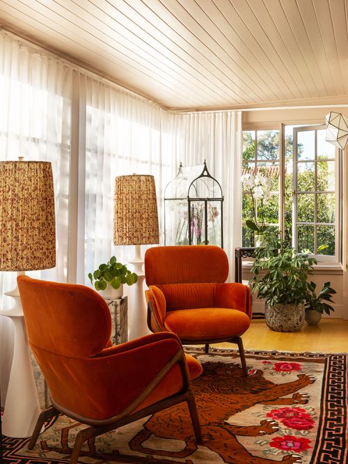 Interior view of living room featuring two orange armchairs, area rug with animal print, and natural light streaming through windows.