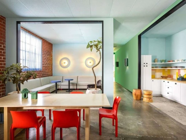 Bright, naturally lit apartment studio with a dining table, a modular sofa, light-blue walls, brick, and a partial view of a modern kitchen.