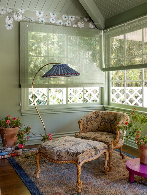 Living room interior with patterned chair and ottoman, floor lamp, area rug, and plants near windows with light green wall paint.