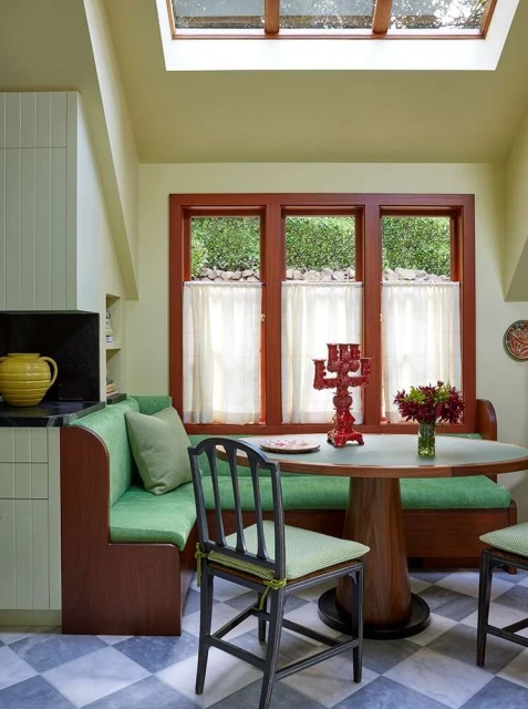Indoor dining nook features a built-in banquette, round table with flowers, chairs, and checkered tile flooring under a skylight.