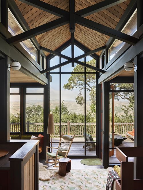 Interior view of a modern living room with floor-to-ceiling windows, a rocking chair, and a view of a wooded landscape.