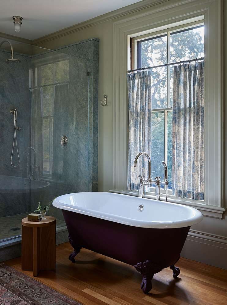 Clawfoot bathtub in a bathroom with wood floors, marble shower, and patterned window curtains bringing natural light indoors.