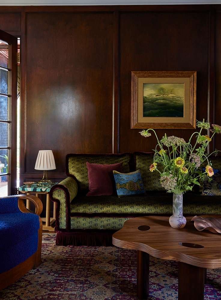 Living room interior with a green patterned sofa, floral arrangement, wood coffee table, patterned rug, and dark wood wall panels.