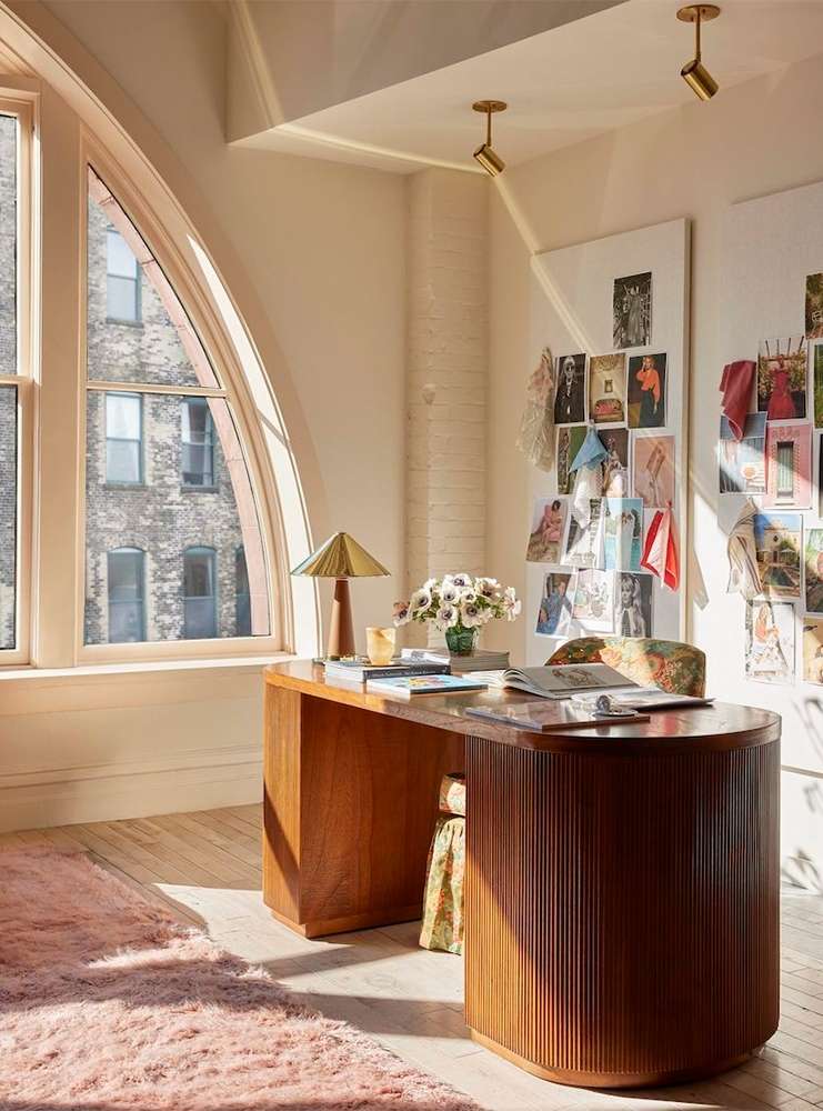 Home office setup with a wooden desk, floral accents, a modern lamp, and natural light filtering through a large arched window.