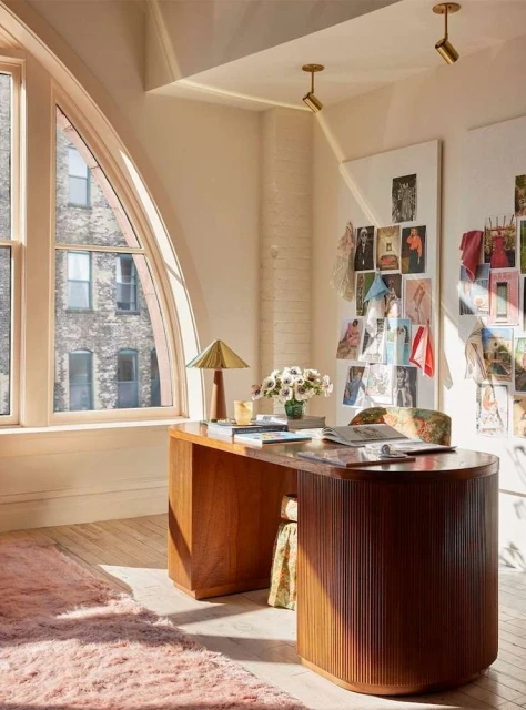 Home office setup with a wooden desk, floral accents, a modern lamp, and natural light filtering through a large arched window.