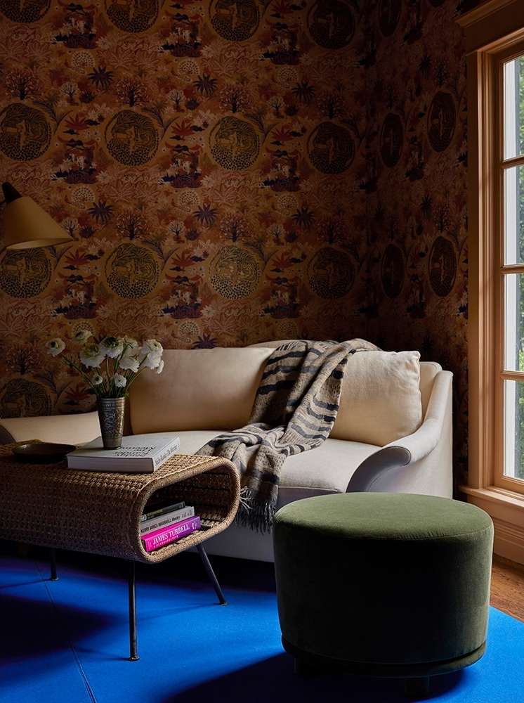 Living room interior with cream sofa, patterned brown wallpaper, woven table, green ottoman, and a blue carpet in natural light.