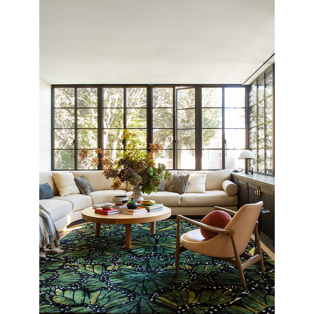 Living room interior with a beige sectional sofa, round wood coffee table, and an armchair, complemented by a green butterfly-patterned rug.