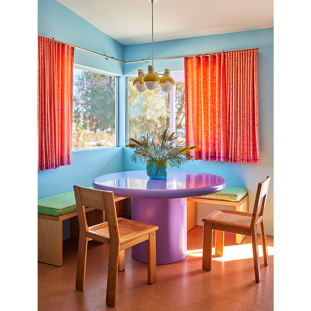 Dining room with a round lavender table, four light chandelier, orange curtains and benches with bright green cushions near windows.