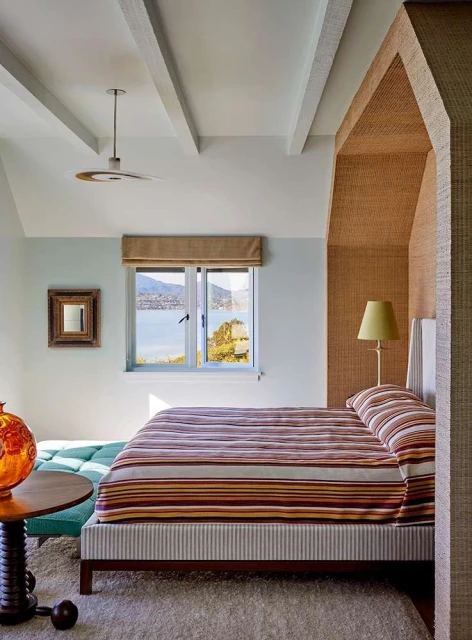Bedroom interior featuring a striped bed, an ocean view through a window, and a vintage-style orange glass lamp on a side table.