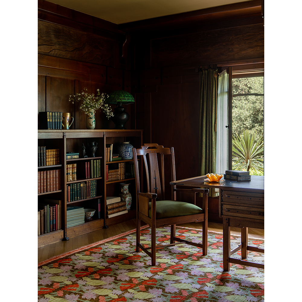 Dark wood interior with a desk, chair, books, flowers, and a green lamp creates a cozy and classic home office scene.
