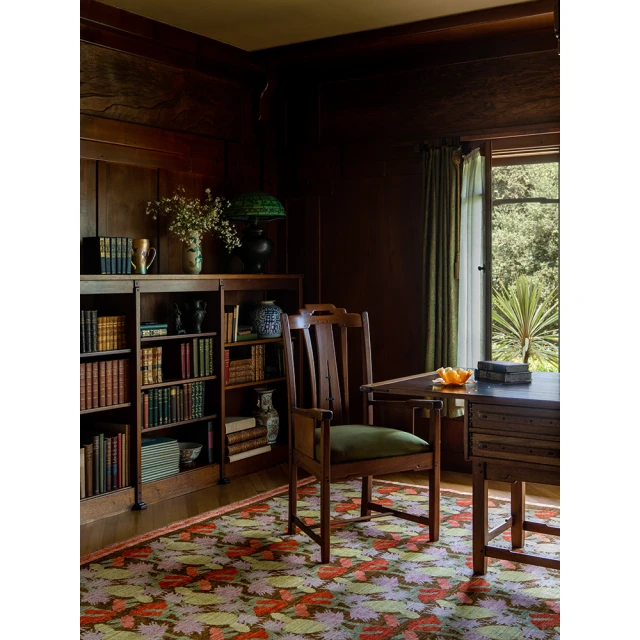 Dark wood interior with a desk, chair, books, flowers, and a green lamp creates a cozy and classic home office scene.
