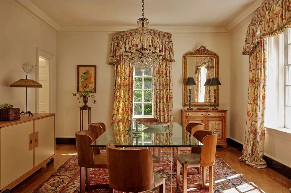 Interior view of a dining room with a glass-top table, wooden chairs, a chandelier, and floral curtains, creating a warm, inviting atmosphere.