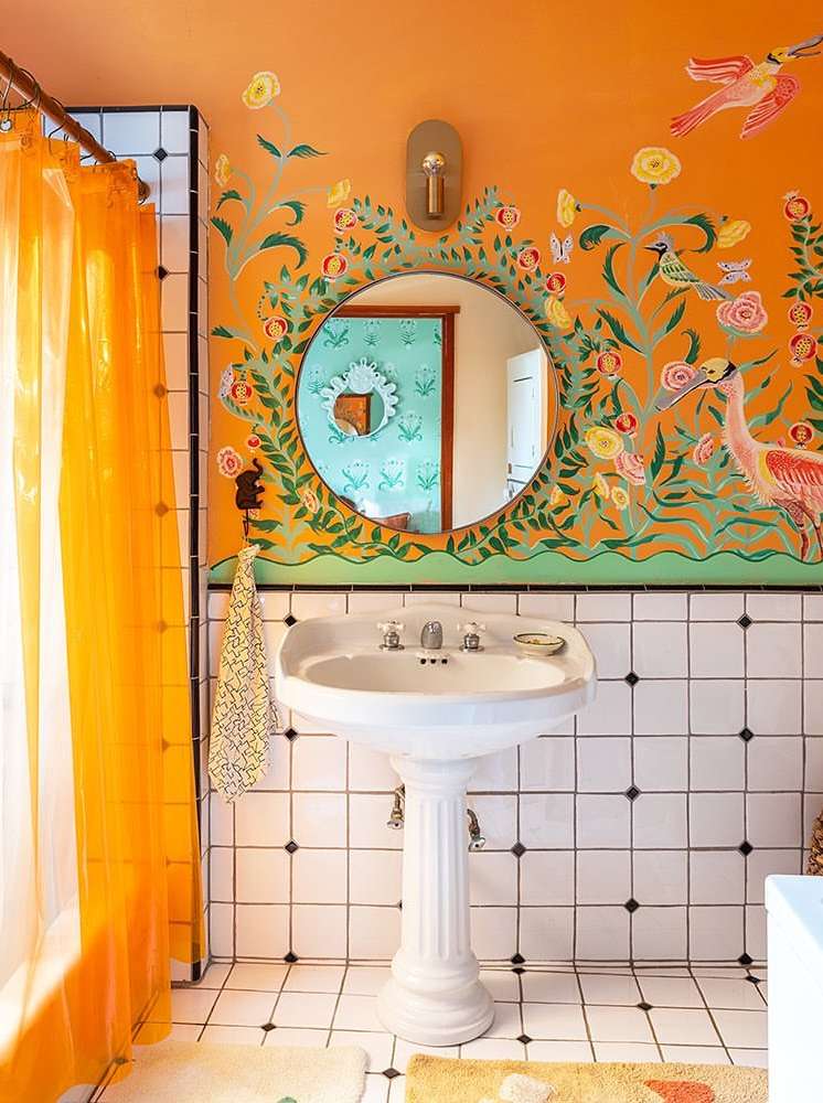 Bathroom interior featuring a pedestal sink, floral wall art, and bright orange shower curtain in a white-tiled room.