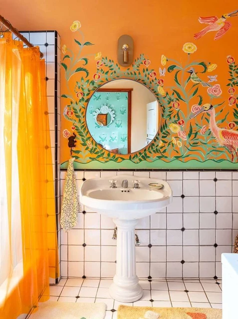 Bathroom interior featuring a pedestal sink, floral wall art, and bright orange shower curtain in a white-tiled room.