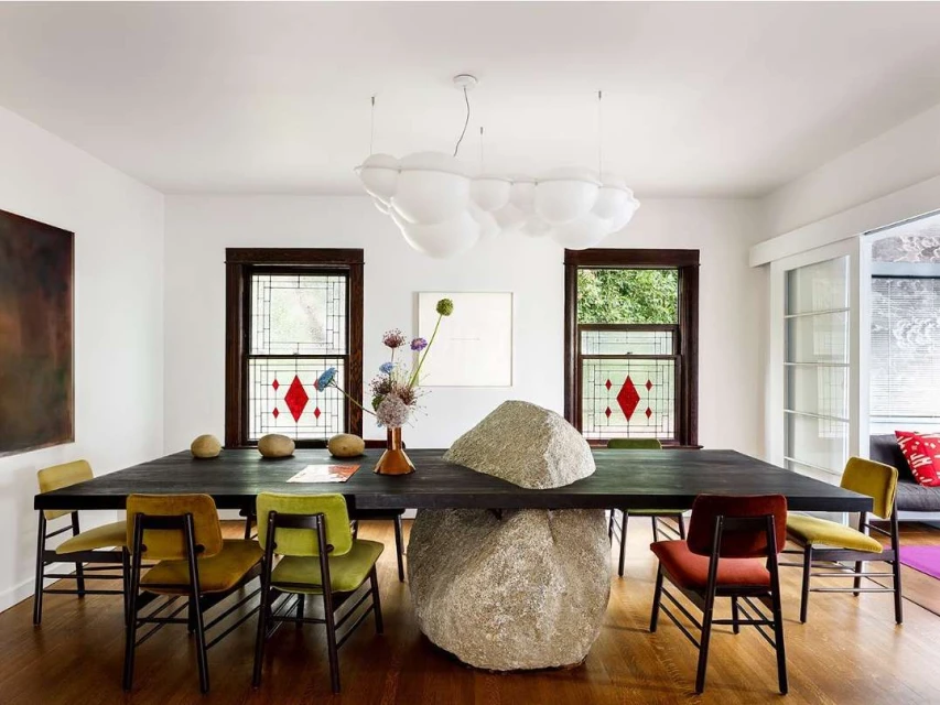 Interior space with a large dark wooden dining table, stone base, colorful chairs, modern light fixture, and natural light from windows.