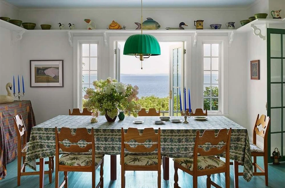 Dining room interior with a large patterned tablecloth, wooden chairs, and ocean view through windows, illuminated by a green pendant light.
