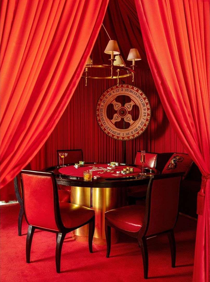 Opulent red casino room with a round poker table, red leather chairs, a roulette wheel, and plush carpeting for a high-stakes game.