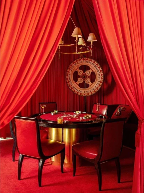 Opulent red casino room with a round poker table, red leather chairs, a roulette wheel, and plush carpeting for a high-stakes game.