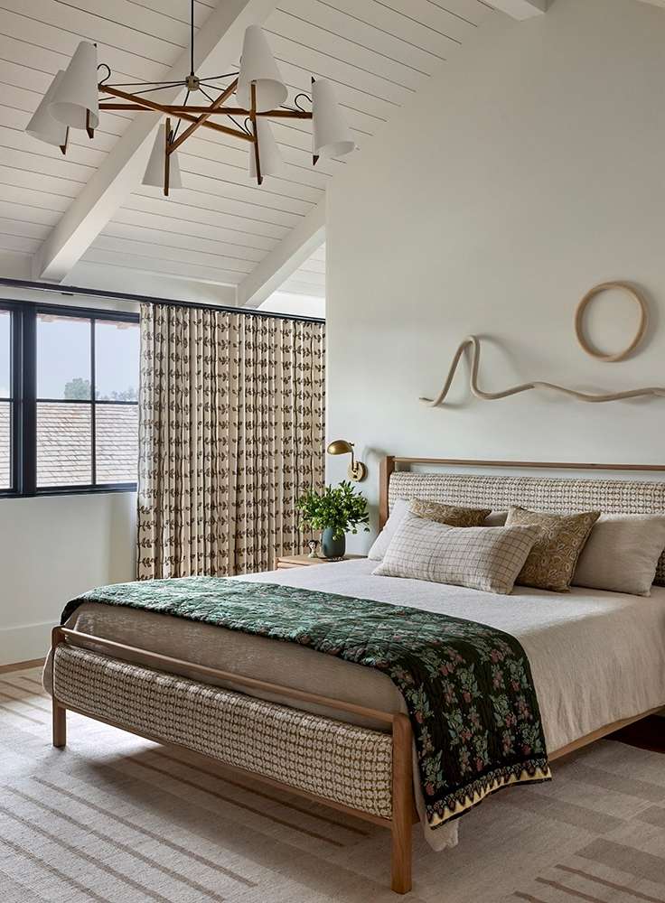 Bedroom interior with a platform bed dressed in neutral linens, a green patterned coverlet, and decorative wall hangings above the headboard.