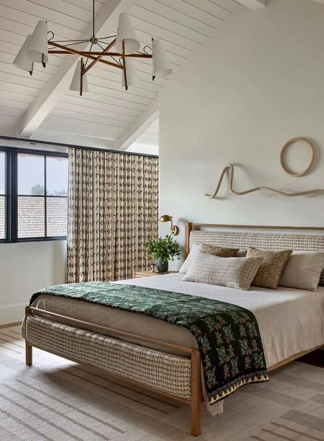 Bedroom interior with a platform bed dressed in neutral linens, a green patterned coverlet, and decorative wall hangings above the headboard.