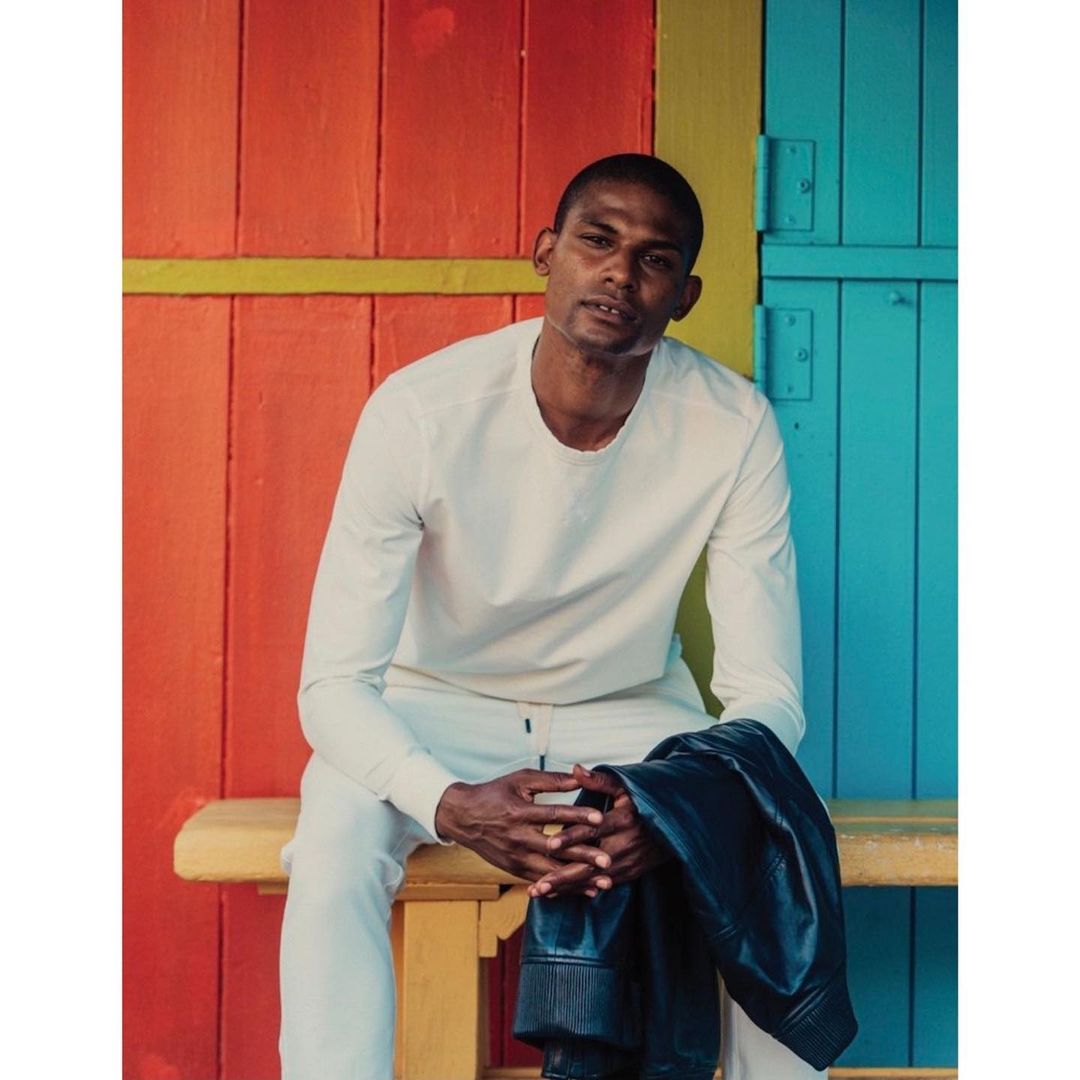 Portrait of an adult male in a white long-sleeve shirt and pants, seated outdoors against a brightly colored wall with a jacket.