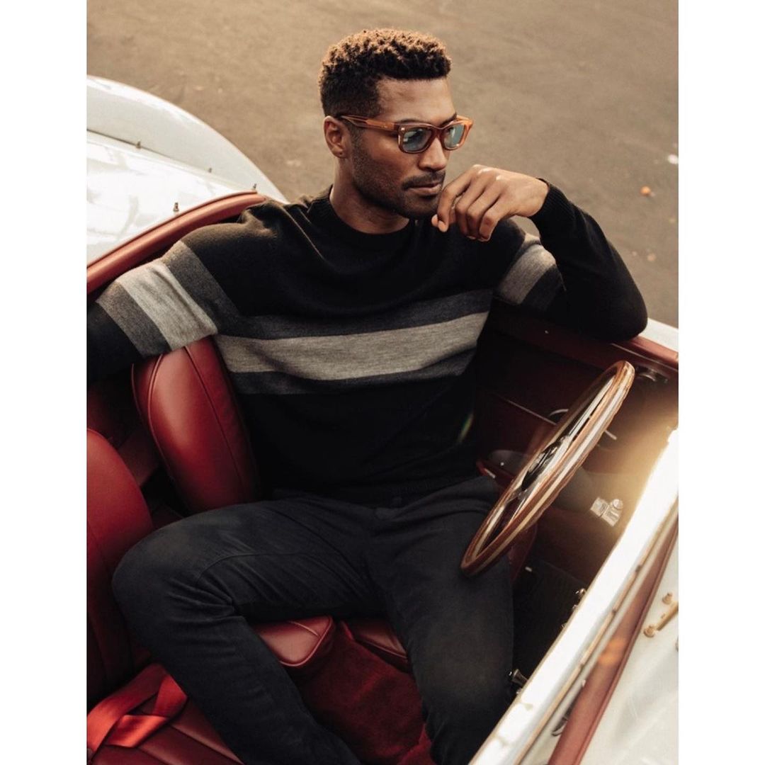 Man in black sweater and black pants sits in a red vintage car wearing sunglasses, looking off to the side with his hand near his chin.