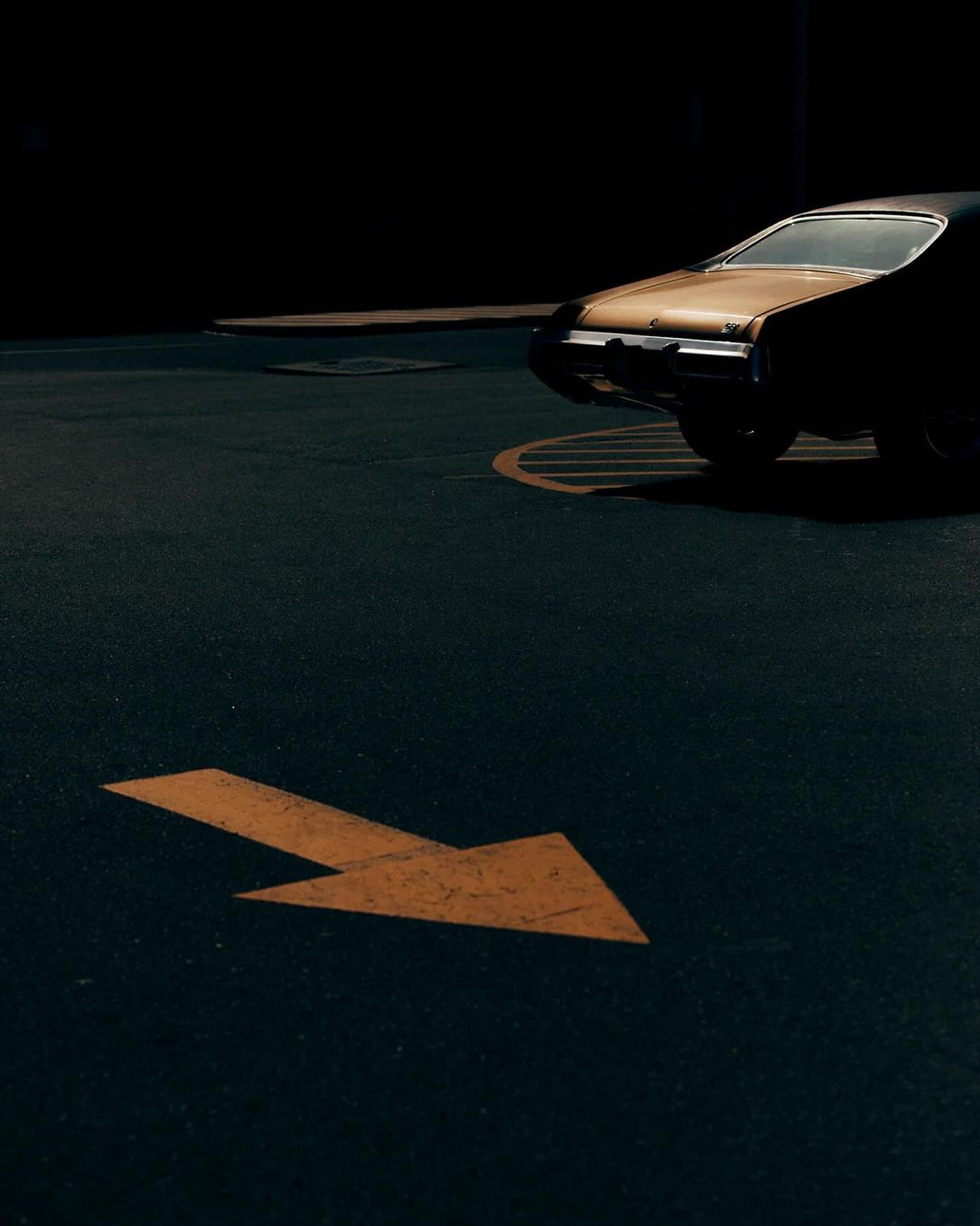 A classic black and gold muscle car parked in a dark parking lot with an orange arrow on the ground pointing to the right.