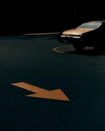 A classic black and gold muscle car parked in a dark parking lot with an orange arrow on the ground pointing to the right.