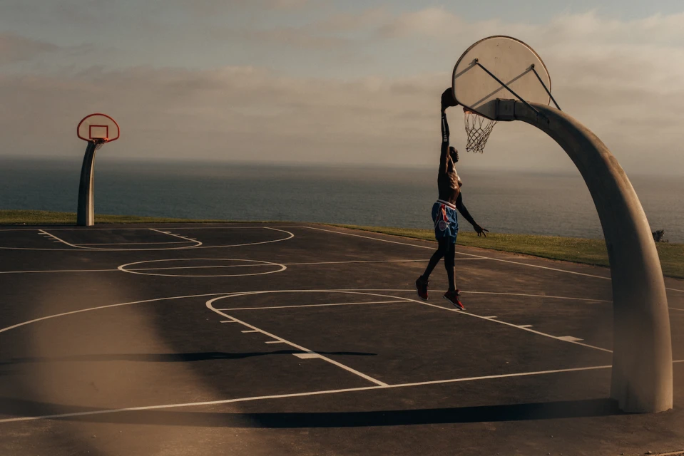 An adult male athlete in sportswear leaps to dunk a basketball on an outdoor court by the ocean in a wide angle shot.