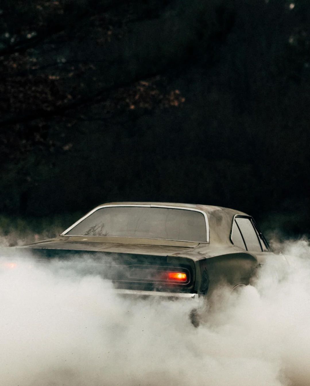 Dark, vintage muscle car emitting smoke in an outdoor environment, captured from a rear view, with dark moody lighting.