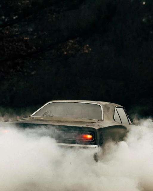 Dark, vintage muscle car emitting smoke in an outdoor environment, captured from a rear view, with dark moody lighting.
