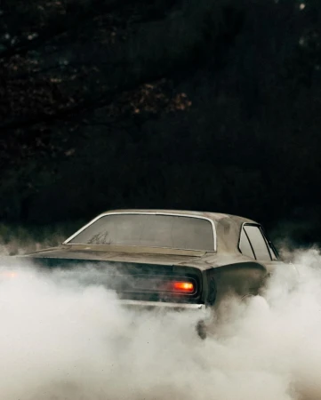 Dark, vintage muscle car emitting smoke in an outdoor environment, captured from a rear view, with dark moody lighting.