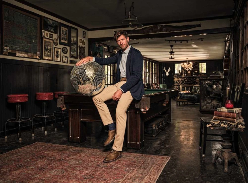 Adult male in a blue jacket sits on a billiard table holding a disco ball in an interior living space with wood paneling and vintage decor.