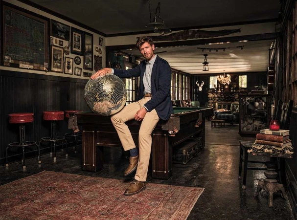 Adult male in a blue jacket sits on a billiard table holding a disco ball in an interior living space with wood paneling and vintage decor.