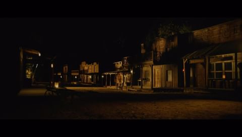Old Western town buildings with wood facades and windows at night, dimly lit by lanterns and street lights, creating a vintage scene.