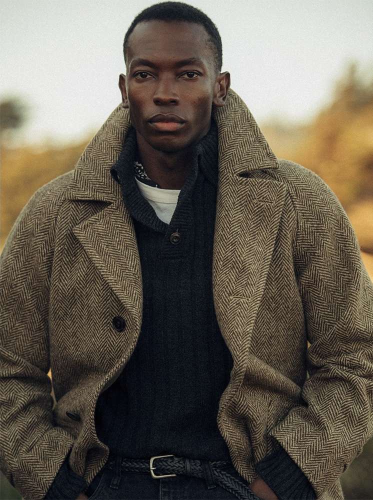 Portrait of a handsome Black man in a herringbone overcoat and sweater, set against an outdoor background with a neutral color palette.