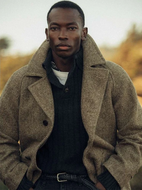 Portrait of a handsome Black man in a herringbone overcoat and sweater, set against an outdoor background with a neutral color palette.