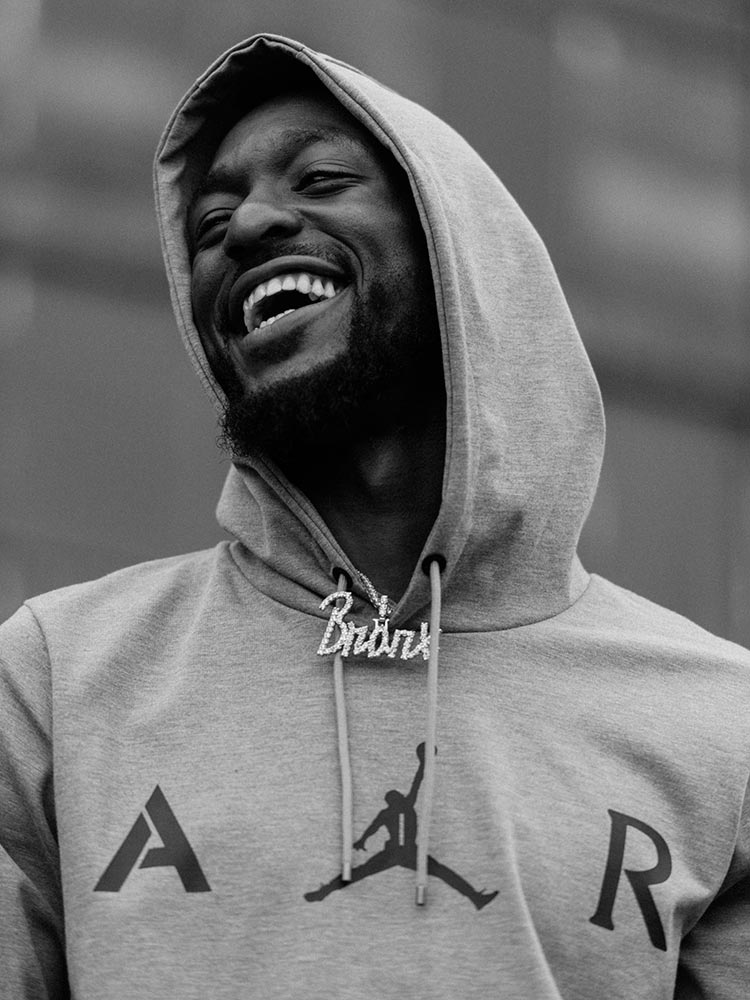 Black and white portrait of a smiling man in a grey hoodie with a custom chain necklace, photographed by Nicholas Maggio.