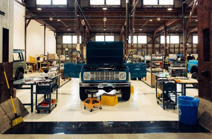Classic turquoise Bronco car in a large garage with its hood open. Car restoration and maintenance is taking place in the well-lit space.